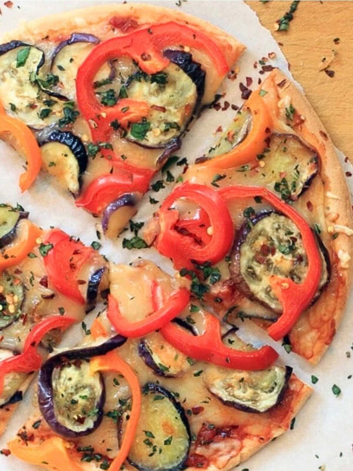 Cheddar cheese pizza topped with eggplant, bell peppers, and herbs on white parchment paper.