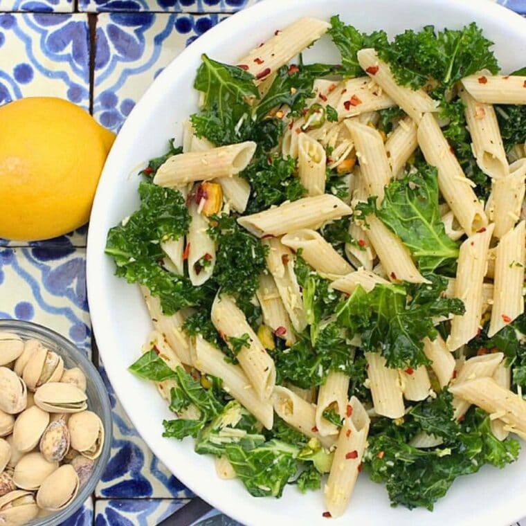 White bowl with pasta, kale, lemon, pistachio nuts, and red pepper flakes.