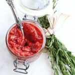 White background with an overhead photo of a jar of strawberry sauce with a spoon sticking out and a bundle of fresh tarragon on the side.