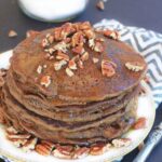 Closeup shot of a white plate with a stack of gingerbread pancakes topped with pecans and maple syrup.