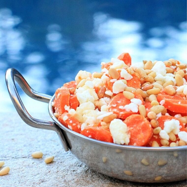 Silver bowl with steamed carrots topped with feta cheese and pine nuts.
