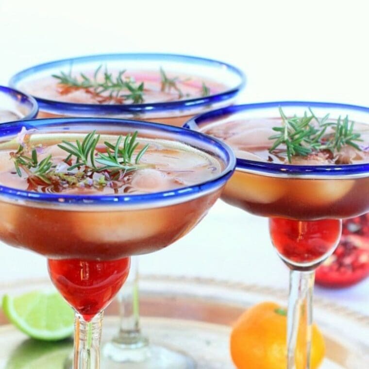 3 pomegranate margaritas garnished with fresh rosemary in blue rimmed glasses.
