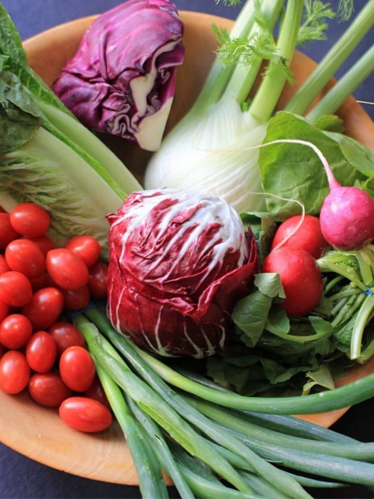 Wooden bowl filled with vegetables: endive, cabbage, leeks, tomatoes, and fennel.