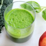 Overhead photo of a glass of green juice with kale, apples, and basil in the background.