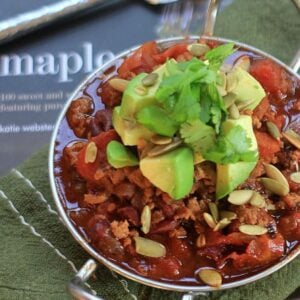 Turkey chili in a silver bowl, topped with avocado, cilantro, and pumpkin seeds.