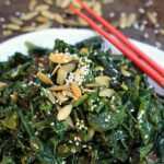Pile of sauted kale and Swiss chard topped with pumpkin seeds and sesame seeds. Red chopsticks in the background.