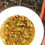 White bowl with vegan split pea soup. Fresh herbs, carrots, and split peas in the background.