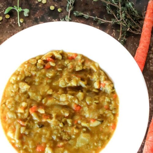 White bowl with vegan split pea soup. Fresh herbs, carrots, and split peas in the background.