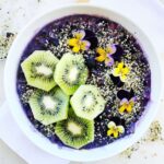 Vegan blueberry smoothie bowl topped with kiwi fruit, hemp seeds, and viola flowers.