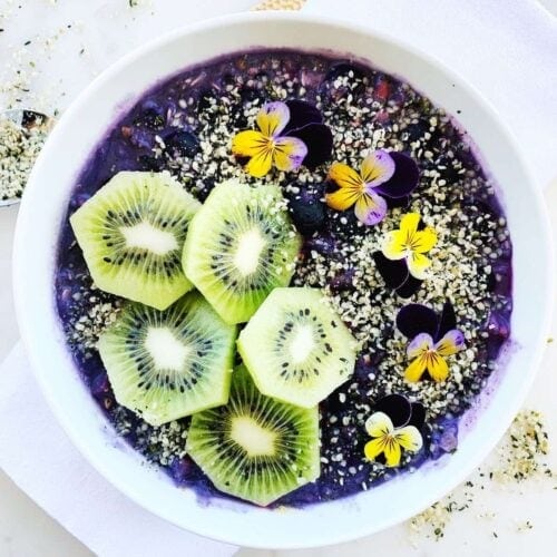 Vegan blueberry smoothie bowl topped with kiwi fruit, hemp seeds, and viola flowers.