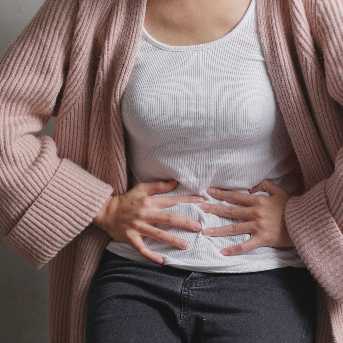 Photo of a woman in a pink sweater and white tank top holding her belly that's in pain from SIBO symptoms.