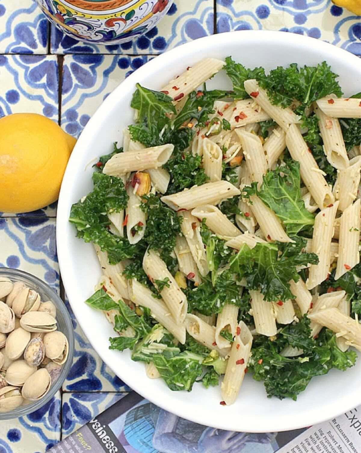 Kale pasta salad in a white bowl with pistachio nuts and a lemon beside it.