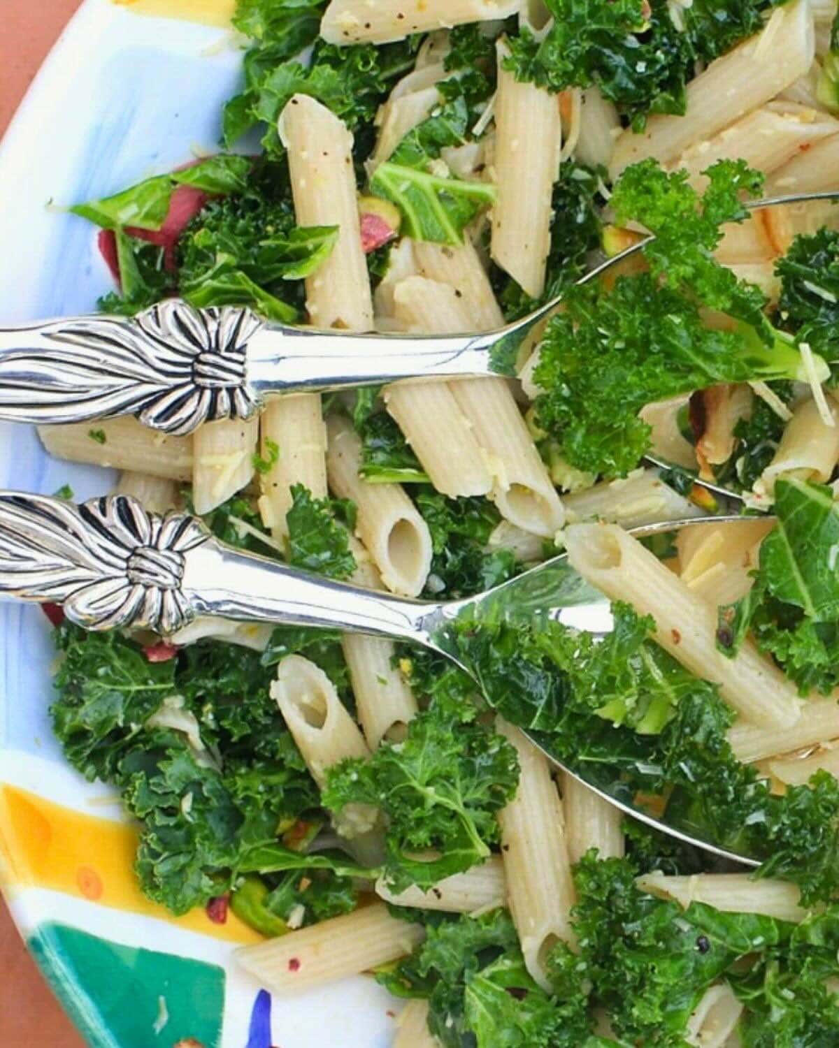 Colorful serving bowl with pasta, kale, and silver serving spoons.