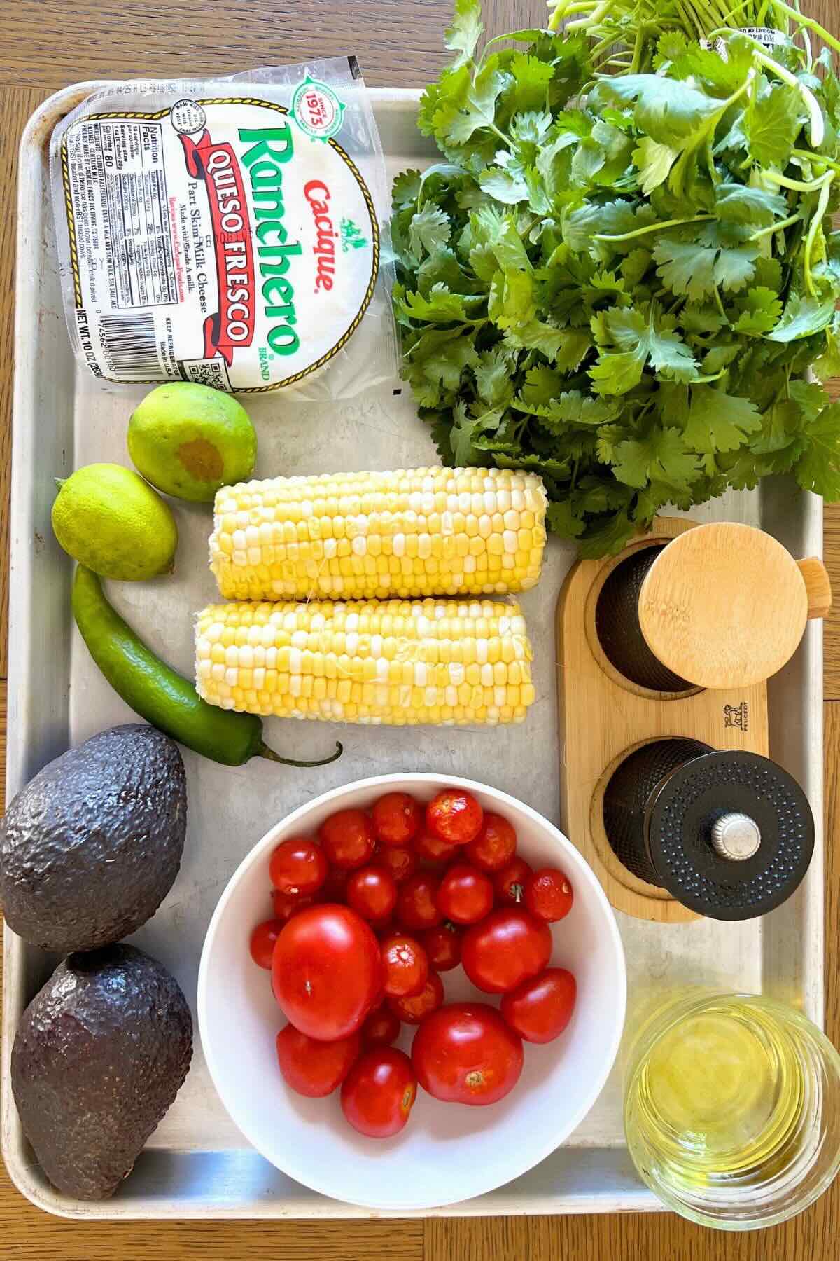 Metal tray with ingredients: corn, cherry tomatoes, avocado, queso fresco, cilantro, oil, limes, salt and pepper.