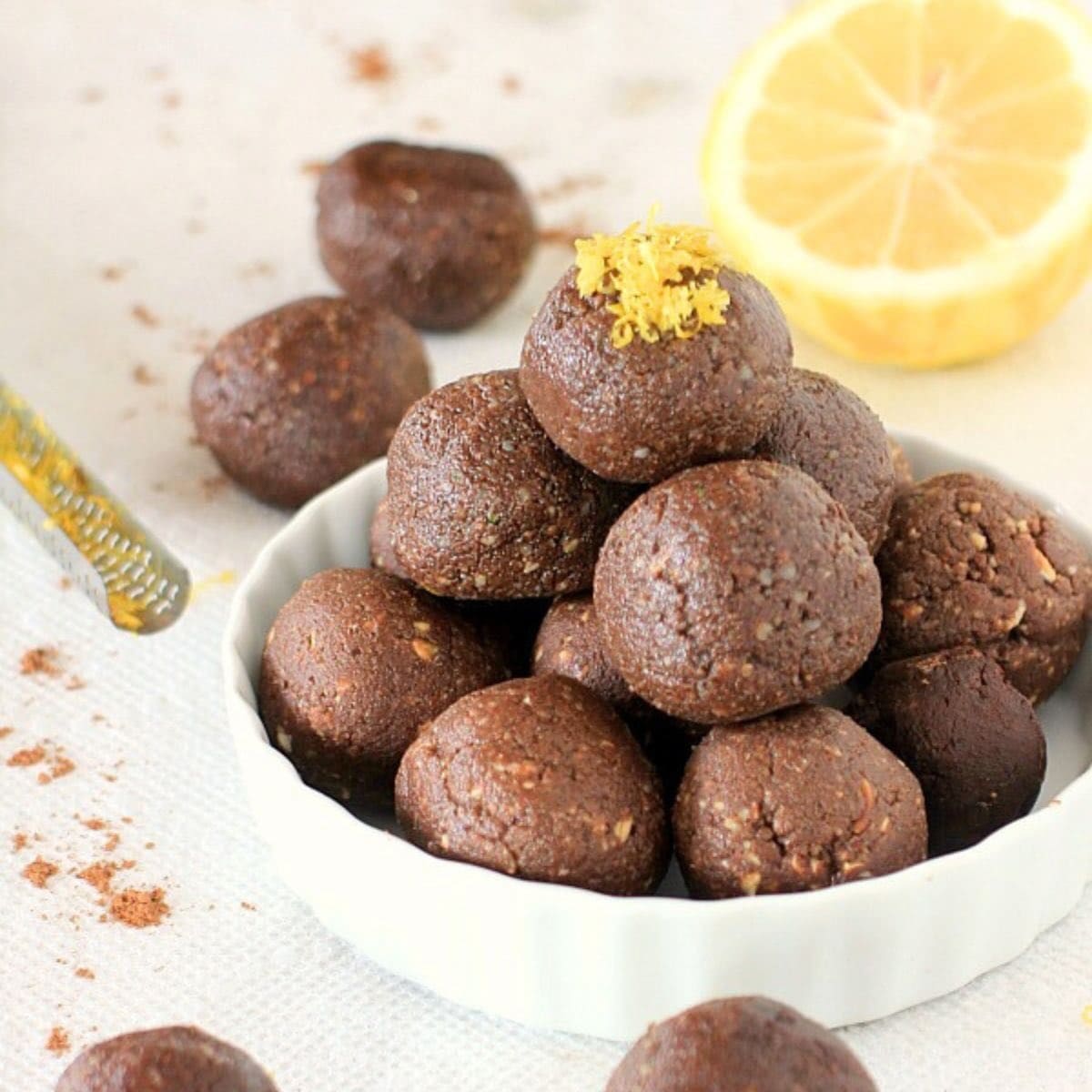 White bowl with cocoa lemon energy bites piled on top. Lemons and lemon zest in the background.