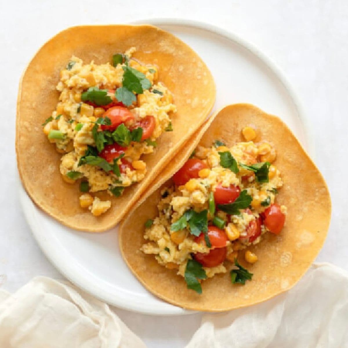 Two breakfast tacos topped with eggs, corn, tomatoes, cilantro, and feta cheese on a white plate.