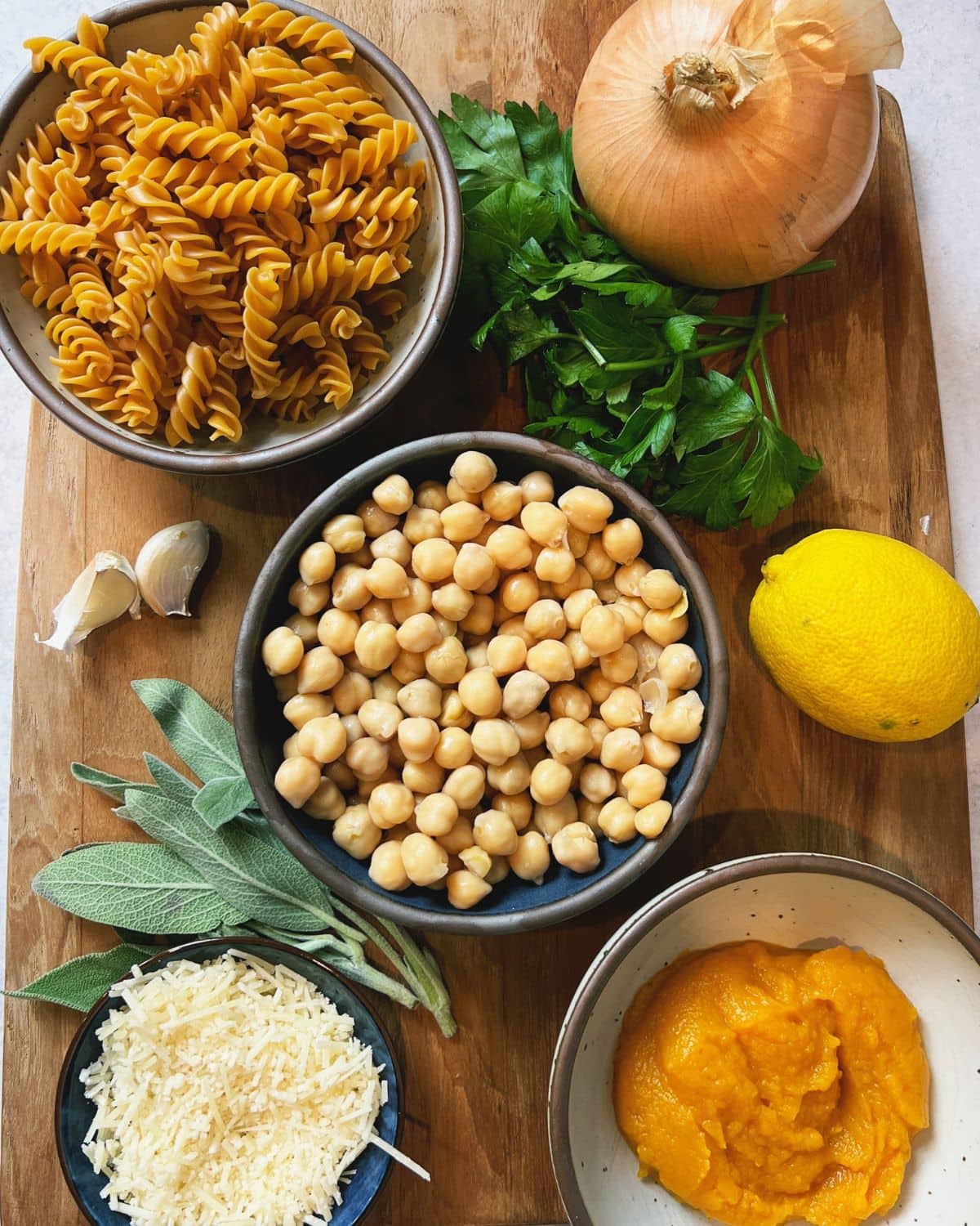Wood cutting board with ingredients: pasta, onion, chickpeas, Parmesan cheese, pumpkin puree, lemon, garlic, fresh parsley, and fresh sage.