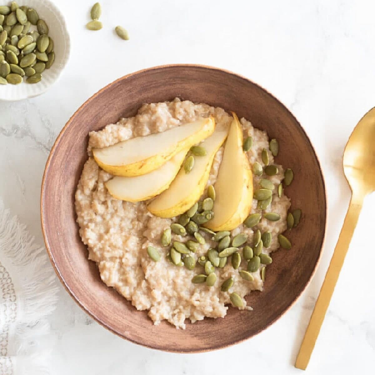 White background with a brown bowl filled with oatmeal topped with sliced pears and pumpkin seeds.