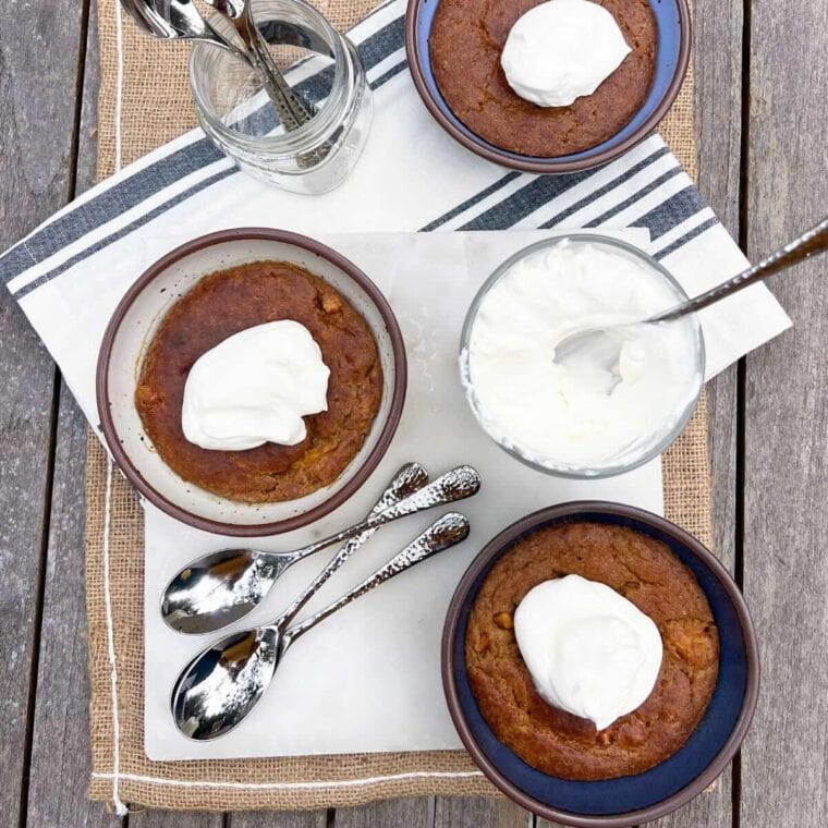 Three bowls of persimmon pudding on a white marble and wood background. Styled with whipped cream and serving spoons.