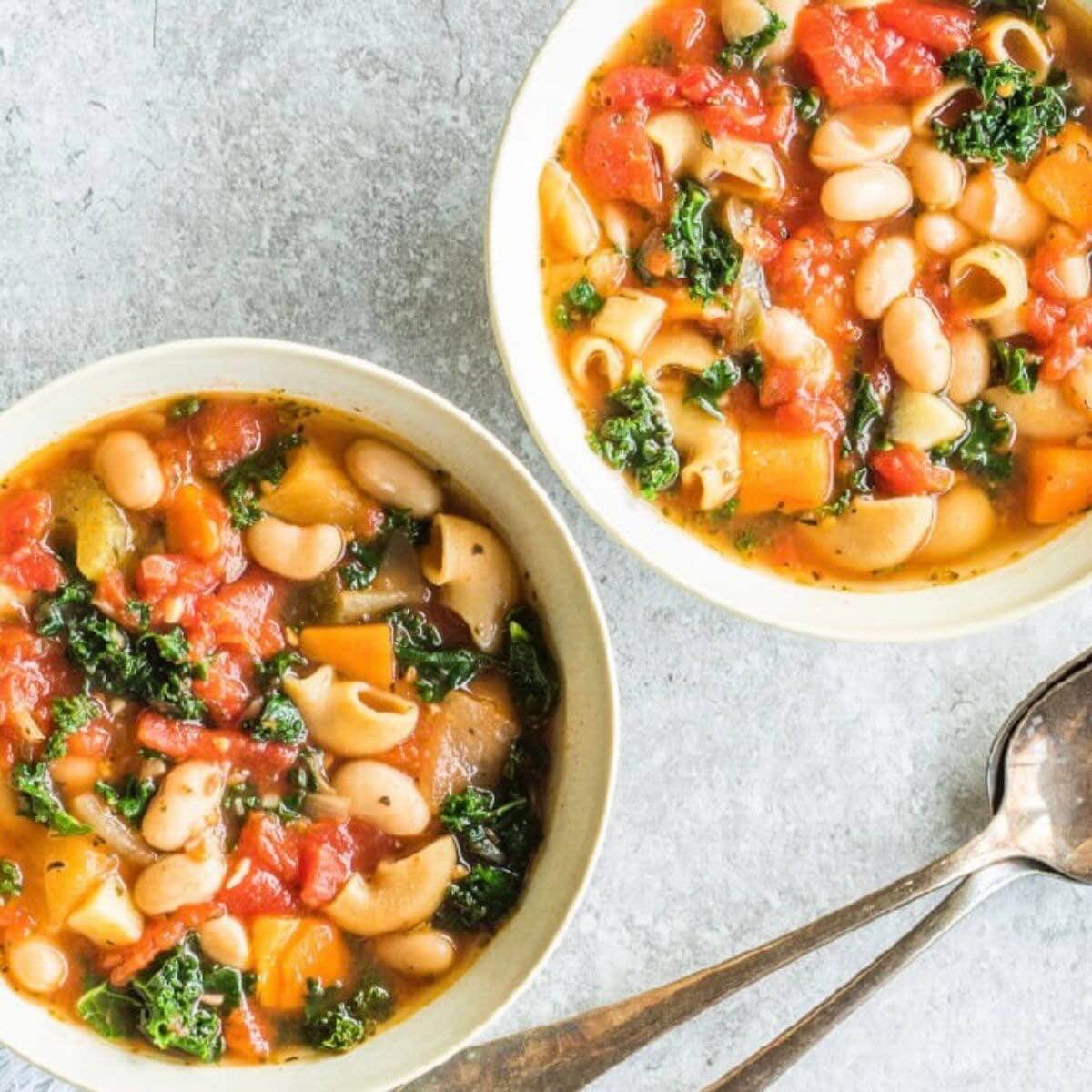 Two white bowls with minestrone soup, and spoons in the background.
