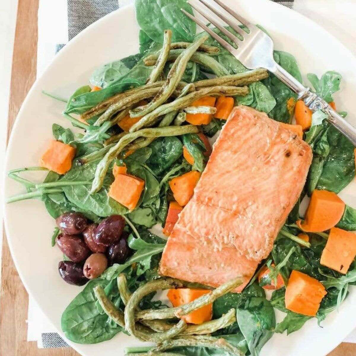 White plate with arugula, kalamata olives, green beans, sweet potatoes, and salmon.