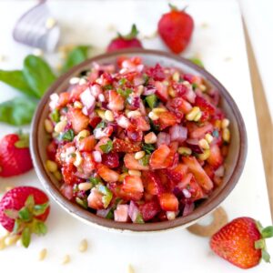 Bowl of strawberry salsa on a white tray with fresh strawberries, pines nuts, and basil in the background.