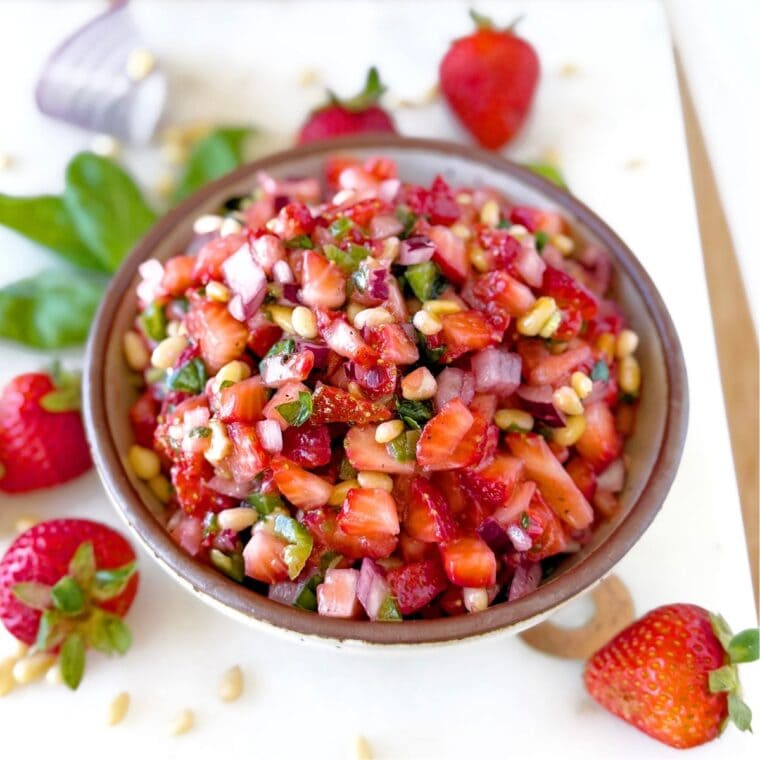 Bowl of strawberry salsa on a white tray with fresh strawberries, pines nuts, and basil in the background.